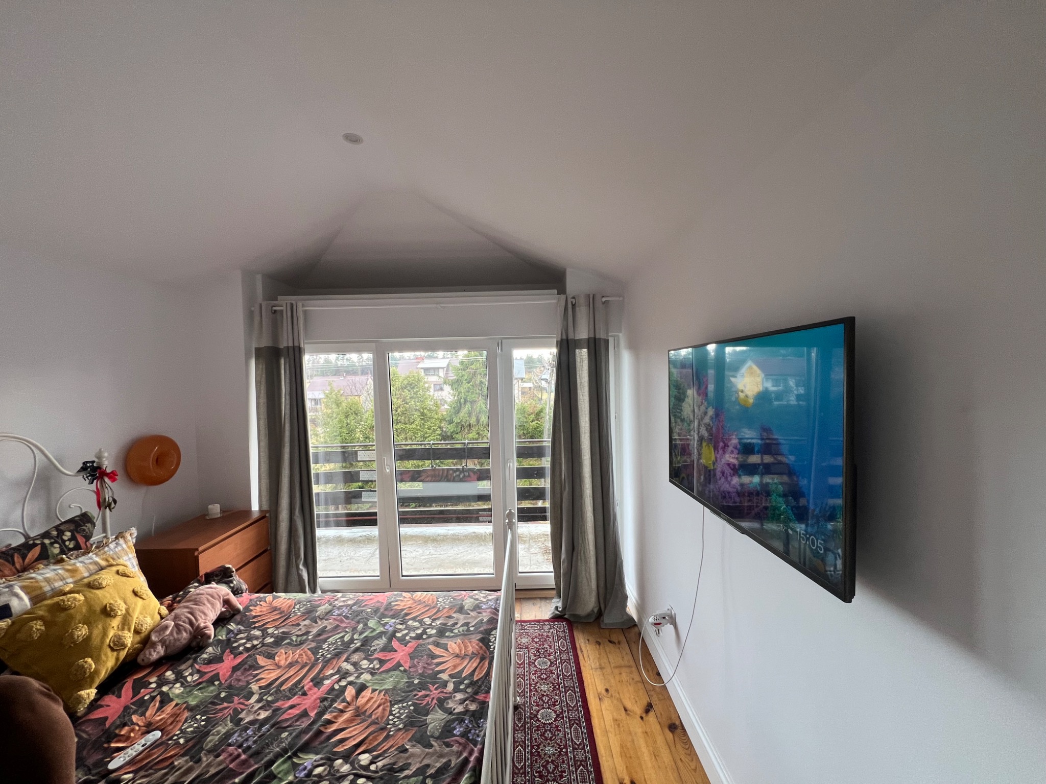 Pokój na poddaszu z łóżkiem, telewizorem na ścianie i wyjściem na balkon. Widoczny fragment dywanu i drewniana podłoga.