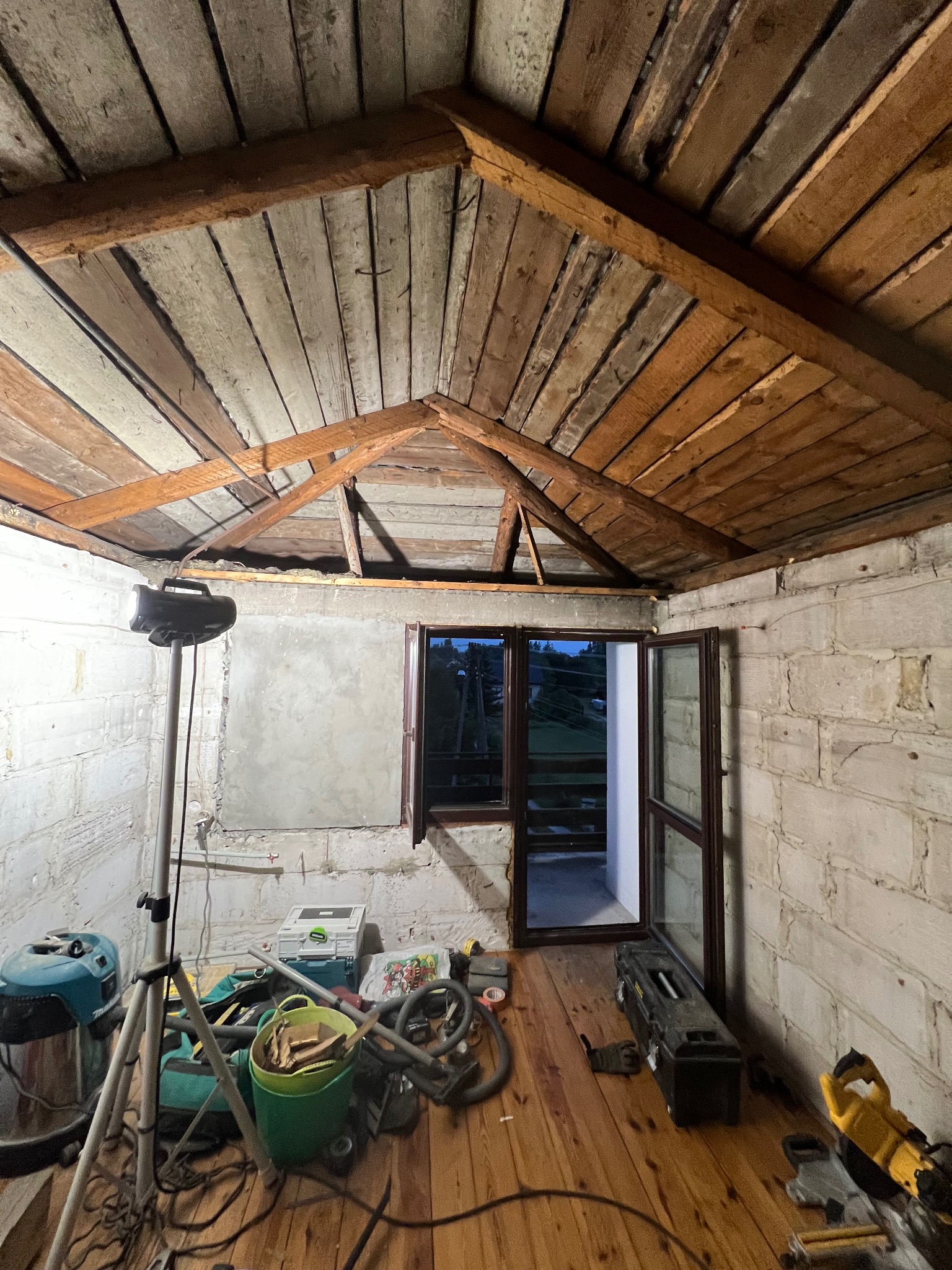 Wnętrze remontowanego pokoju z drewnianym stropem, oknem balkonowym i przygotowaną ścianą do położenia warstwy wykończeniowej, widoczne narzędzia i oświetlenie robocze.