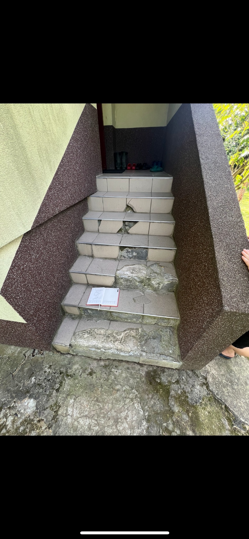 Zniszczone schody wejściowe z popękanymi płytkami, notatnik z rysunkiem leżący na jednym ze stopni, widoczne ślady wilgoci i uszkodzenia na powierzchni stopni.