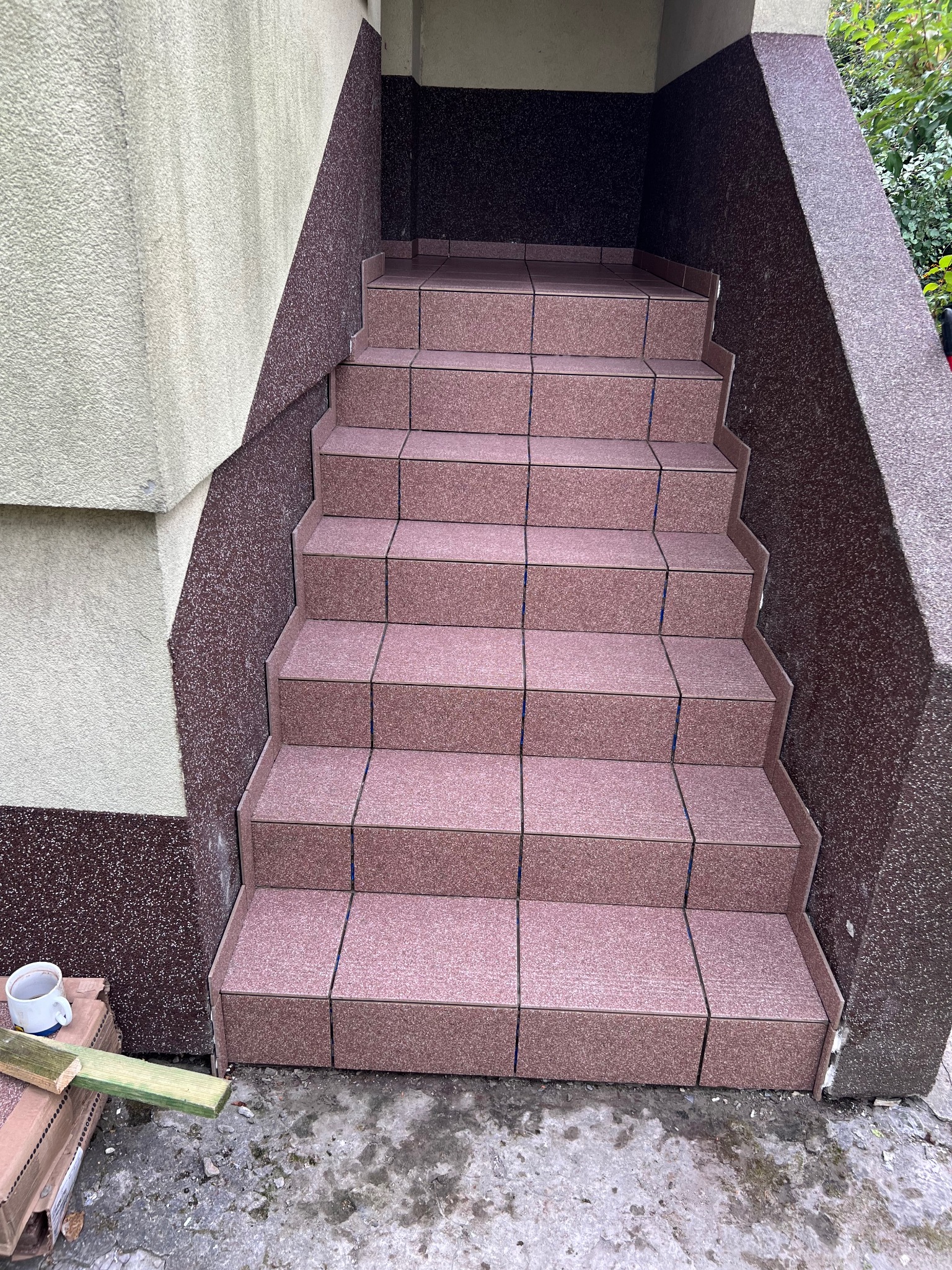 Wyłożone płytkami schody wejściowe do budynku, widoczne niedokończone prace, obok leży kubek i resztki materiałów budowlanych.