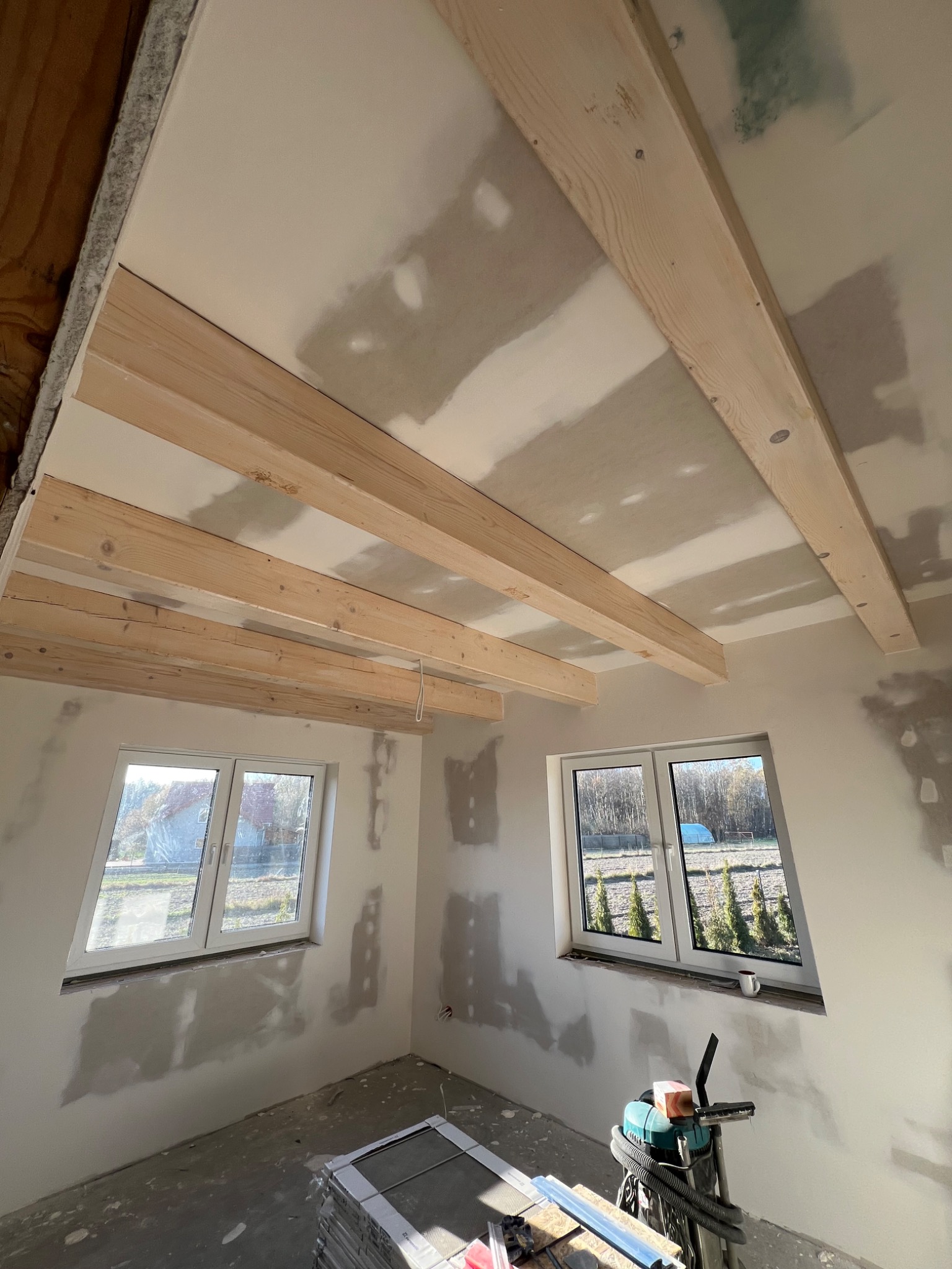 Wnętrze w trakcie prac wykończeniowych: sufit z drewnianymi belkami i częściowo nałożoną warstwą szpachli, ściany z widocznymi śladami przygotowania pod malowanie, dwa okna z widokiem na zewnątrz...