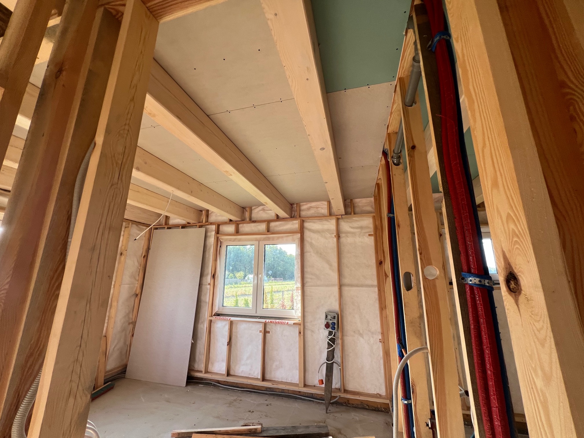 Wnętrze w trakcie budowy, widoczne drewniane ramy ścian, izolacja z wełny mineralnej, częściowo zamontowane płyty gipsowo-kartonowe na suficie, instalacja elektryczna i rury grzewcze w ścianach.