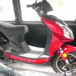 Czerwony skuter marki Romet zabezpieczony pasami wewnątrz furgonetki podczas transportu. Widoczne detale: przednie zawieszenie, opona, logo marki oraz fragment wnętrza pojazdu.
