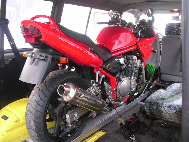 Czerwony motocykl Suzuki Bandit zabezpieczony pasami w przestrzeni ładunkowej busa, przygotowany do transportu. Widoczna rampa załadunkowa i elementy mocujące.