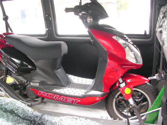 Czerwony skuter marki Romet zabezpieczony pasami wewnątrz furgonetki podczas transportu. Widoczne detale: przednie zawieszenie, opona, logo marki oraz fragment wnętrza pojazdu.