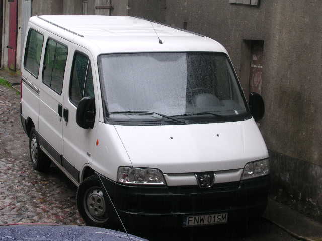 Biały bus marki Peugeot zaparkowany na brukowanej ulicy w wąskiej uliczce, z widoczną tablicą rejestracyjną FNW 01SM.