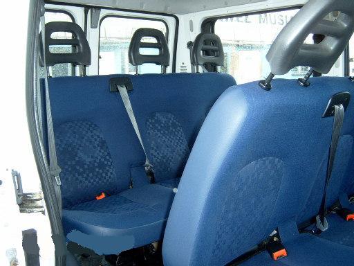 Wnętrze białego busa z trzema rzędami niebieskich foteli z materiałową tapicerką w kratkę, widoczne pasy bezpieczeństwa i zagłówki.