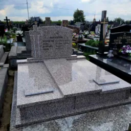 Granitowy nagrobek z jasnoszarego kamienia na cmentarzu, z wygrawerowanymi imionami i datami, otoczony innymi grobami w tle.