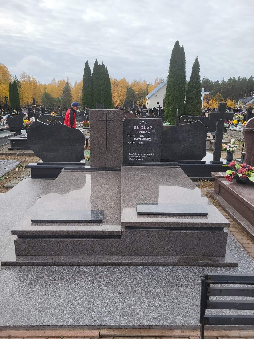Granitowy nagrobek z wygrawerowanym krzyżem i inskrypcjami na cmentarzu, z widoczną sylwetką osoby w czerwonej kurtce w tle.