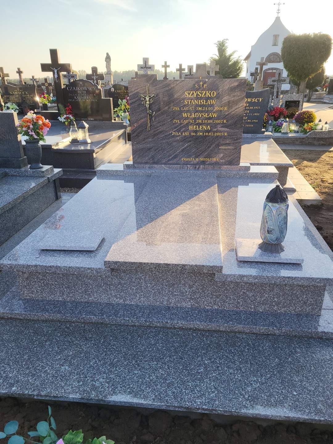 Nowy, wielopoziomowy nagrobek z szarego granitu z inskrypcjami i metalowym krucyfiksem na cmentarzu, widoczny lampion nagrobny i kwiaty na sąsiednich grobach.