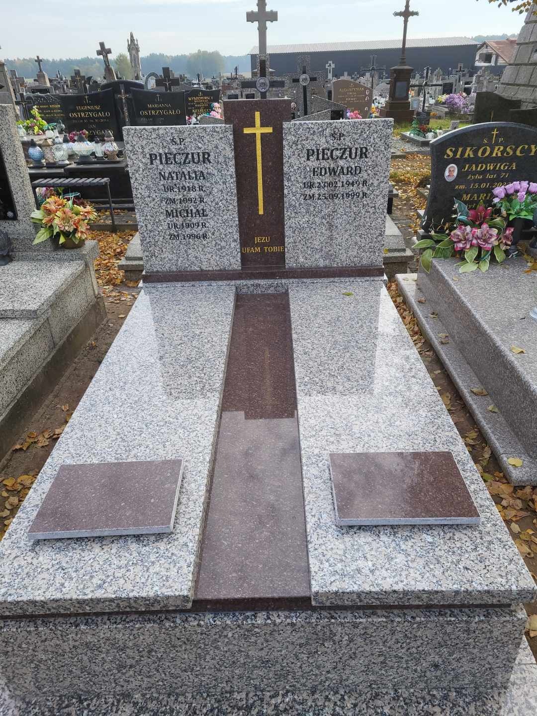 Granitowy nagrobek z wygrawerowanymi imionami i datami, ozdobiony złotym krzyżem, na cmentarzu z widocznymi innymi grobami w tle.