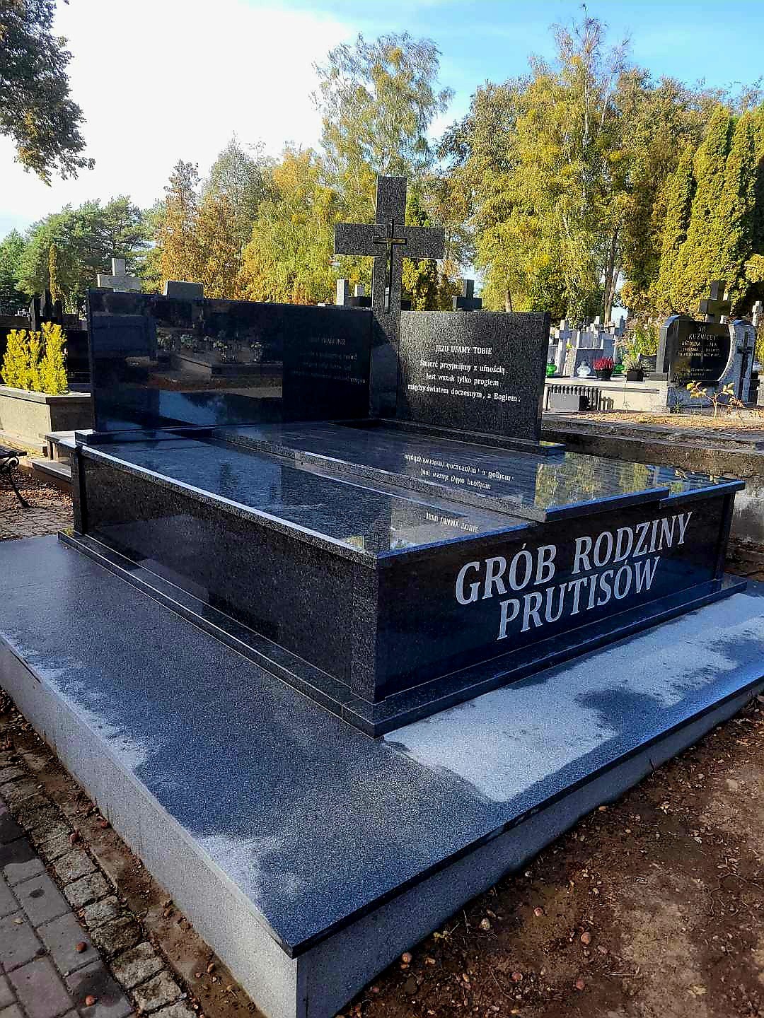 Czarny, polerowany nagrobek rodzinny z granitu z wygrawerowanym napisem 'Grób Rodziny Prutisów' na cmentarzu, otoczony drzewami i innymi nagrobkami.