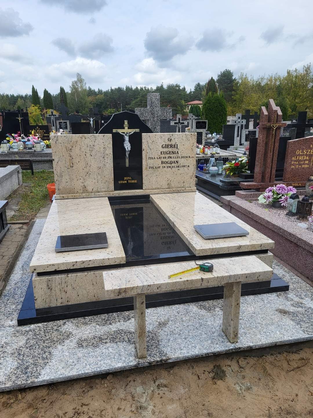 Granitowy nagrobek z czarnymi elementami i krzyżem z Jezusem, na cmentarzu widoczne inne groby, na dolnej płycie leży zwinięta miarka.