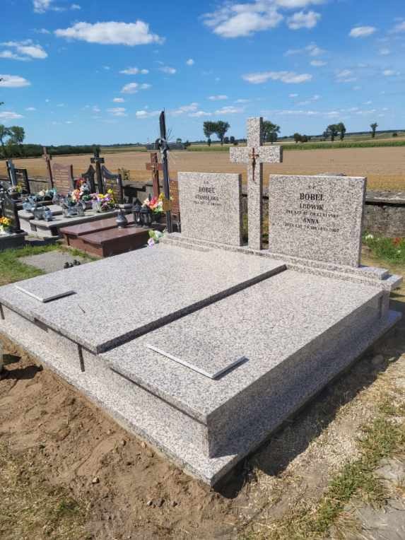 Jasnoszary, dwuosobowy nagrobek z granitu z dwoma tablicami nagrobnymi i krzyżem na cmentarzu, na tle pola i błękitnego nieba z chmurami.