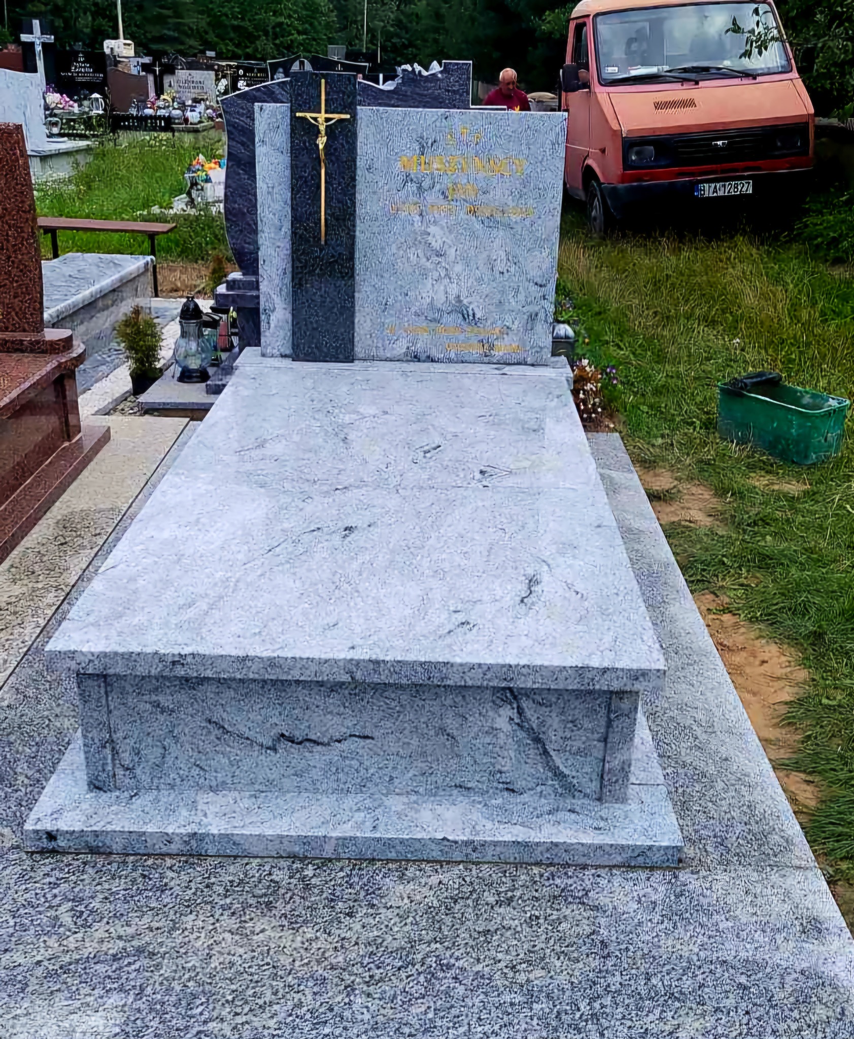 Szary nagrobek z konglomeratu z złotym krzyżem, widoczny na cmentarzu, z pomarańczowym samochodem dostawczym w tle i osobą w czerwonej koszulce