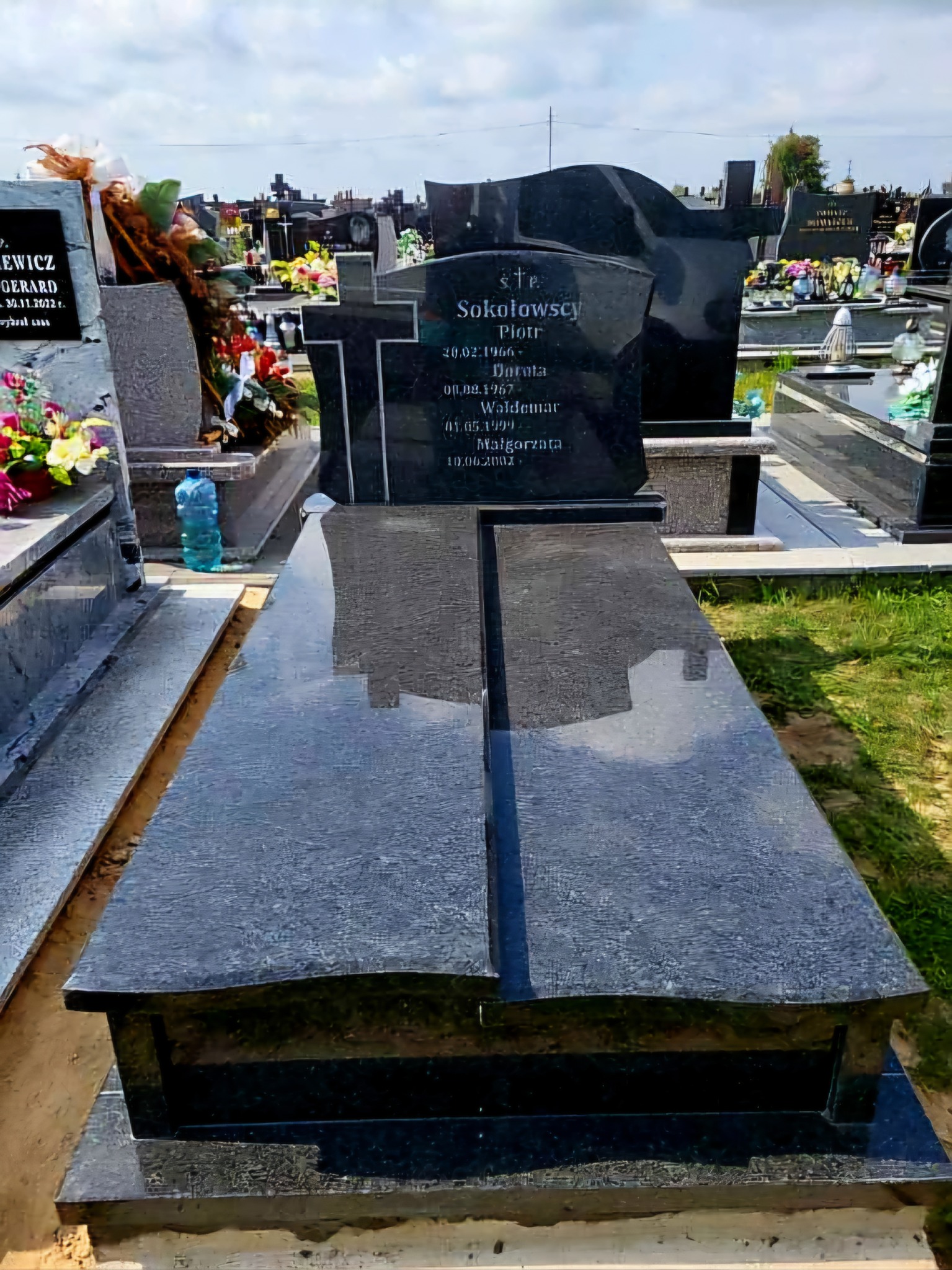 Czarny, podwójny nagrobek z wygrawerowanymi danymi osobowymi na cmentarzu, widok z dołu, w tle inne groby i butelka wody.