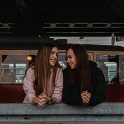 Magdalena Falba - Dwie uśmiechnięte kobiety, jedna w różowej, druga w zielonej bluzie, opierają się o szarą barierkę na tle tramwaju z pasażerami w środku.