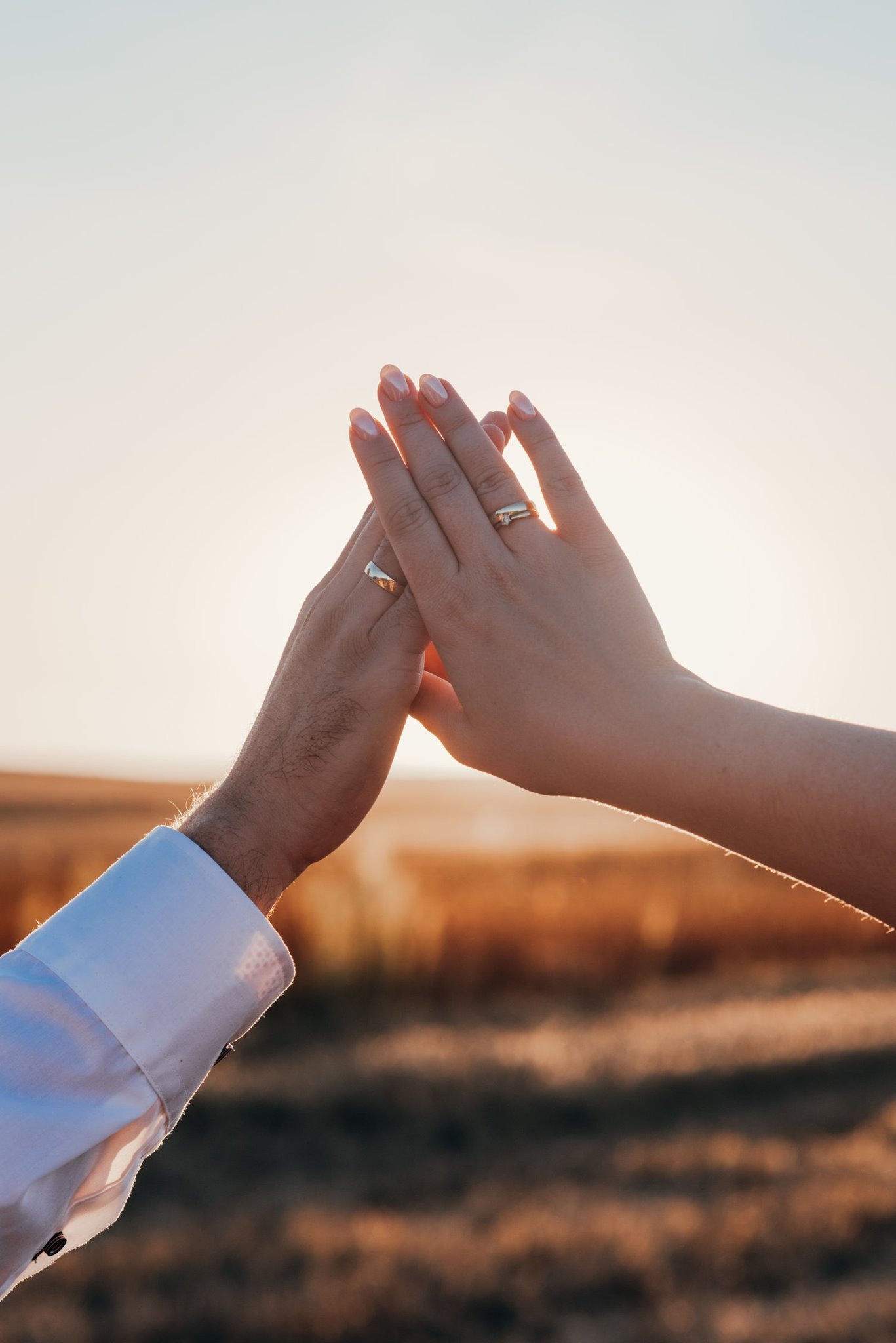 Dłonie pary z obrączkami ślubnymi na tle pola zboża w ciepłym świetle zachodzącego słońca.