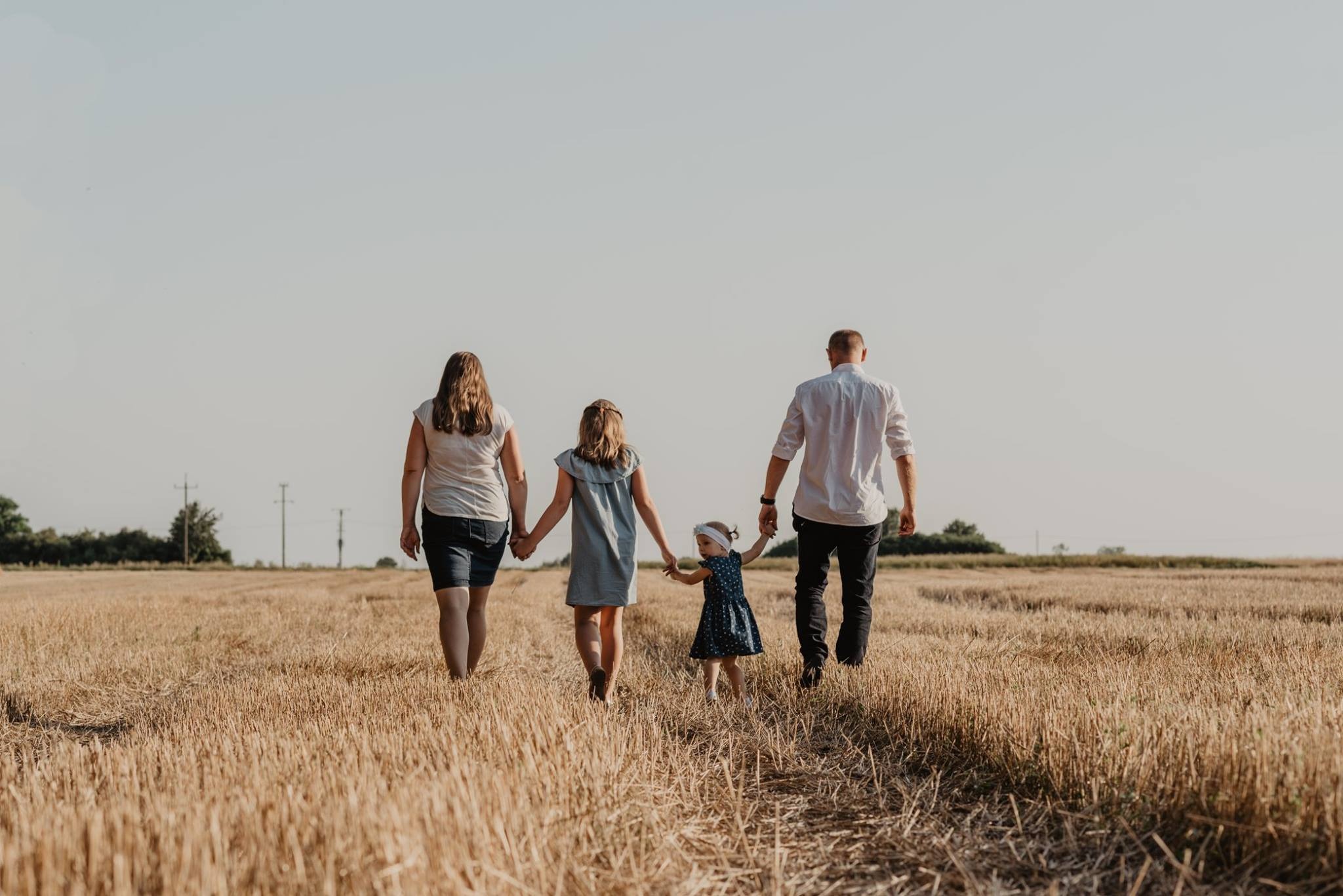 Rodzina z dwójką dzieci, widziana od tyłu, spaceruje po polu ze ściętym zbożem w słoneczny dzień.