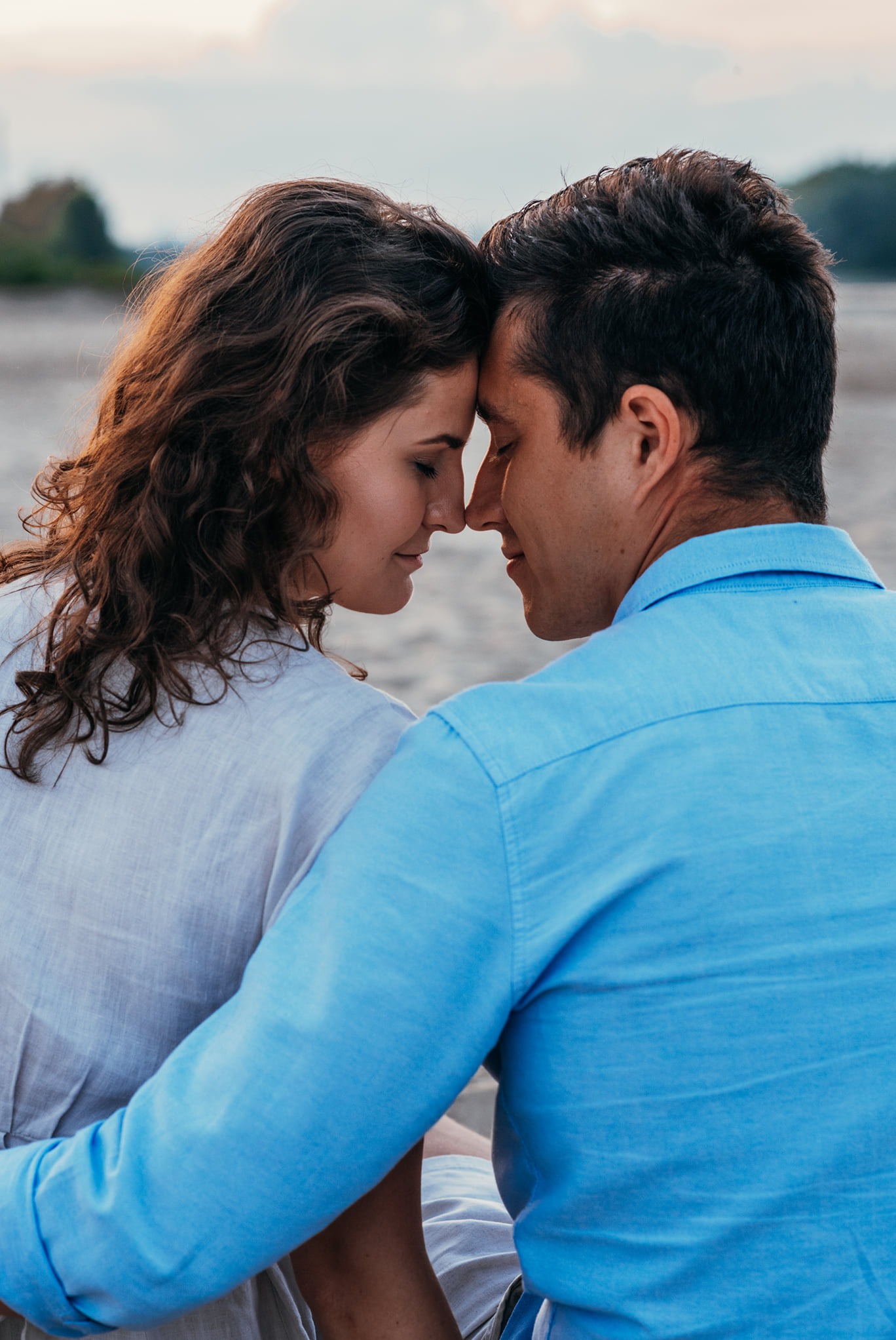 Zakochana para siedzi blisko, dotykając się czołami na plaży o zmierzchu, kobieta z kręconymi włosami w jasnej lnianej koszuli, mężczyzna w błękitnej koszuli z podwiniętymi rękawami.