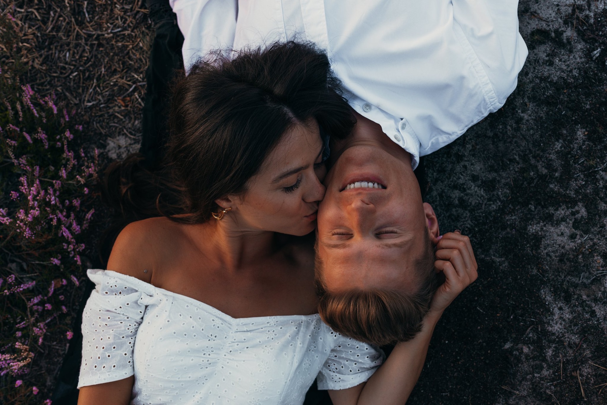 Zakochana para leżąca na ziemi, kobieta całuje mężczyznę w policzek, oboje ubrani na biało, otoczeni wrzosem.