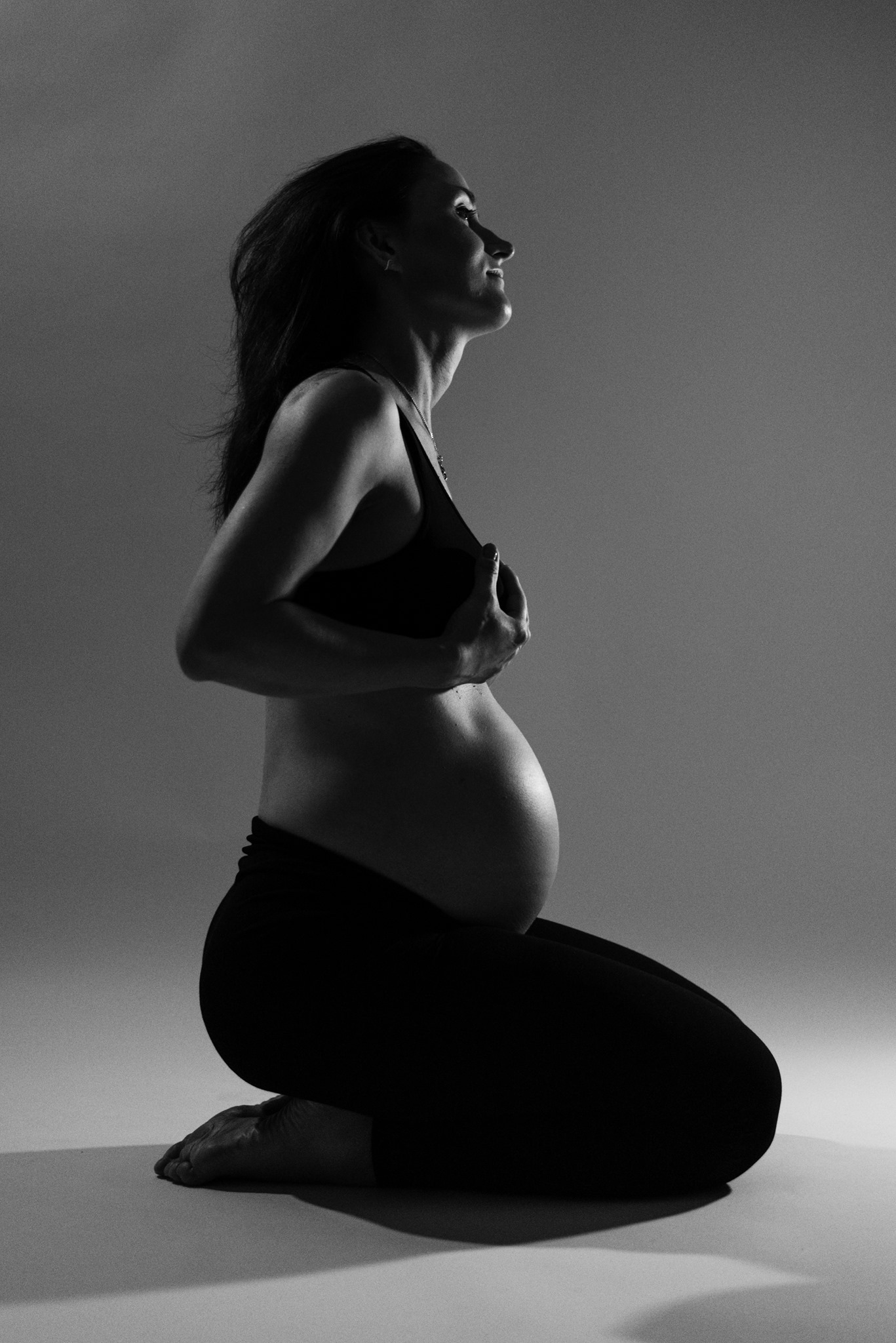 Czarno-biała sylwetka kobiety w ciąży klęczącej na podłodze, z rękami na piersiach, profil twarzy skierowany ku górze, delikatne oświetlenie podkreśla kształt brzucha.