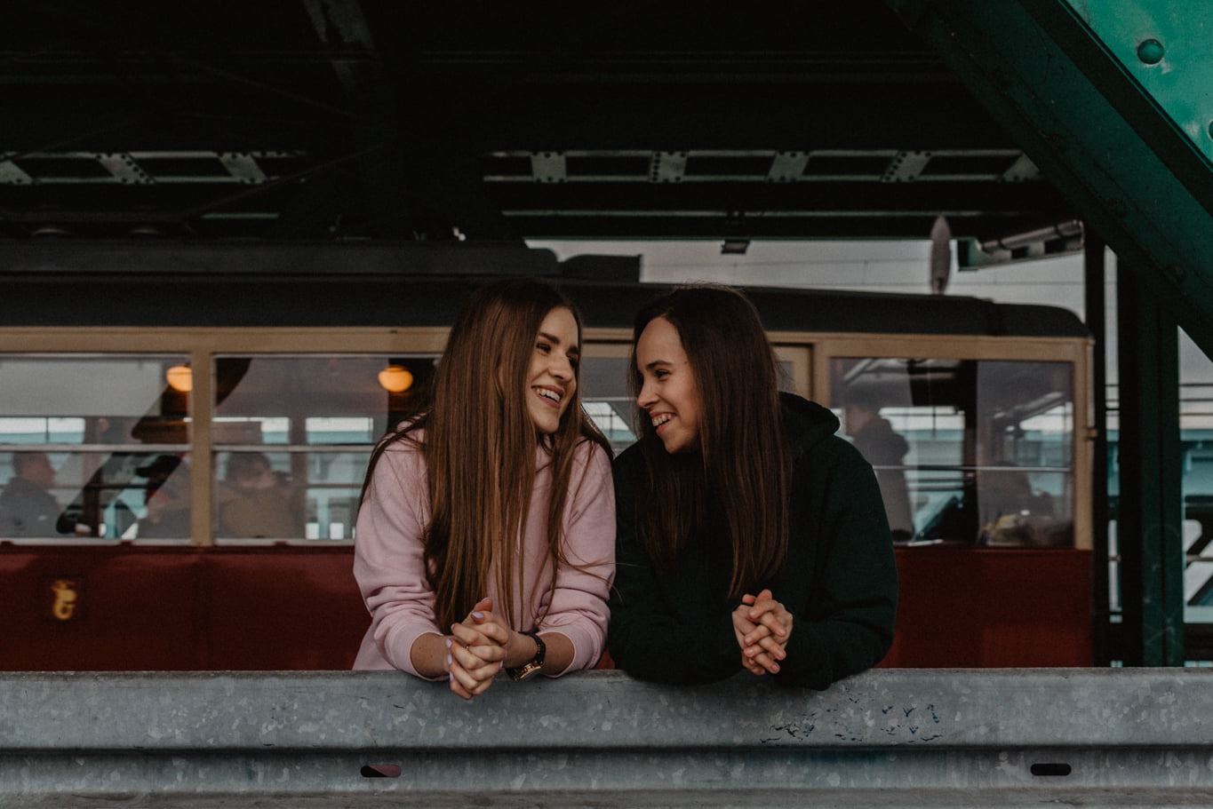 Dwie uśmiechnięte kobiety, jedna w różowej, druga w zielonej bluzie, opierają się o szarą barierkę na tle tramwaju z pasażerami w środku.