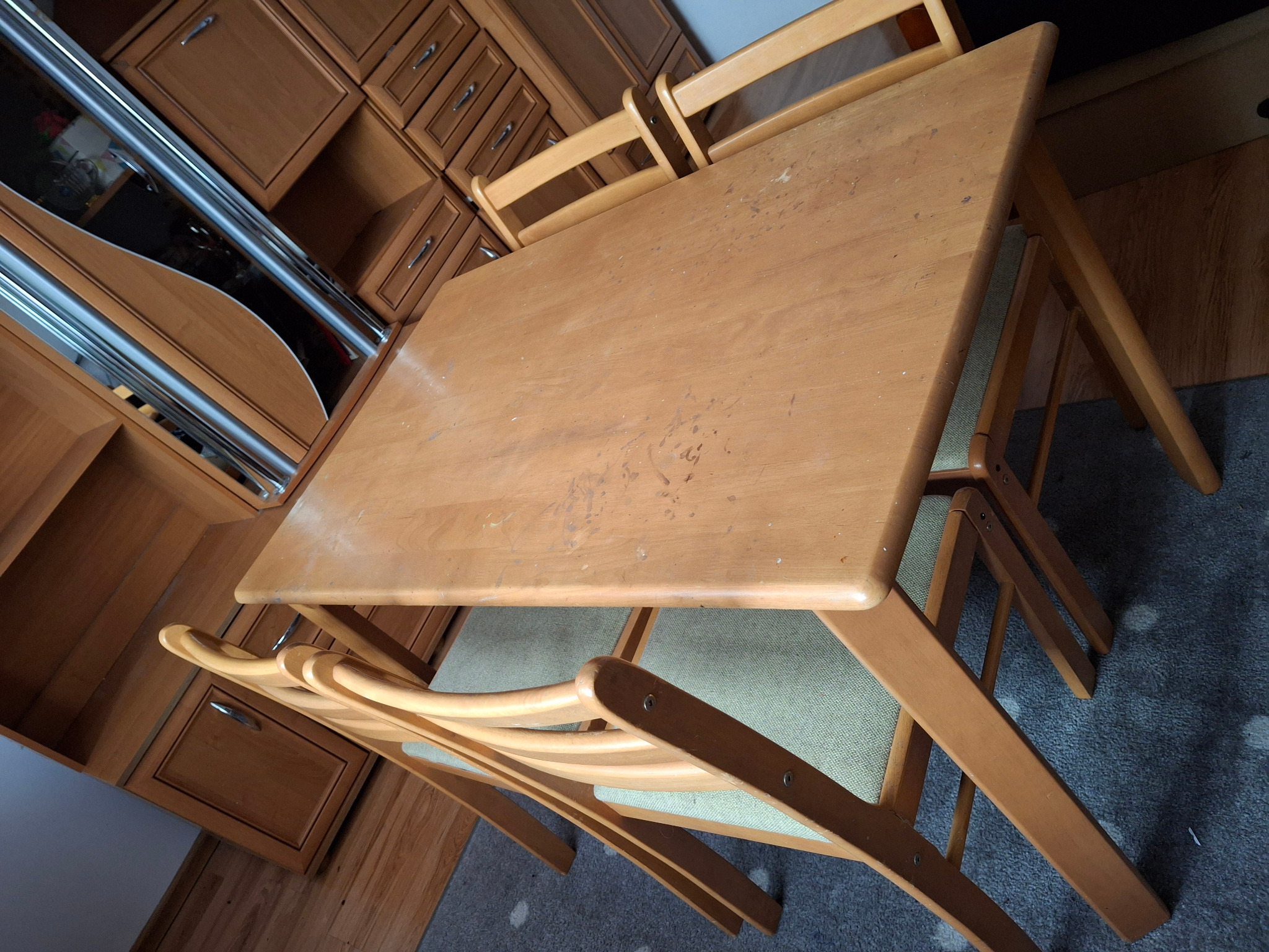 Drewniany stół z krzesłami w pokoju z meblami w zabudowie, widoczne ślady użytkowania na blacie stołu.