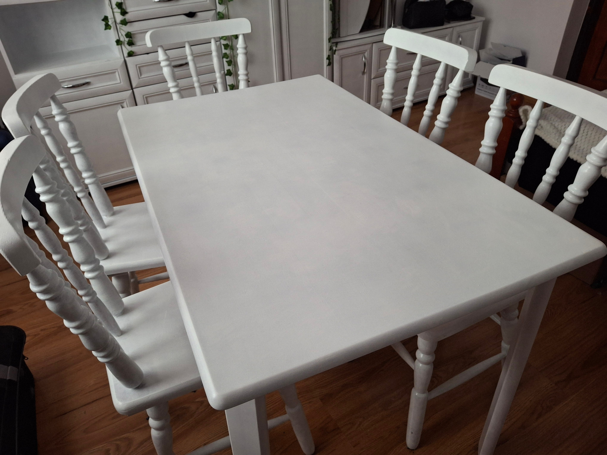 Biały stół z czterema krzesłami w stylu rustykalnym, po renowacji, widoczne ślady pędzla na blacie, w tle białe meble z dekoracją roślinną.