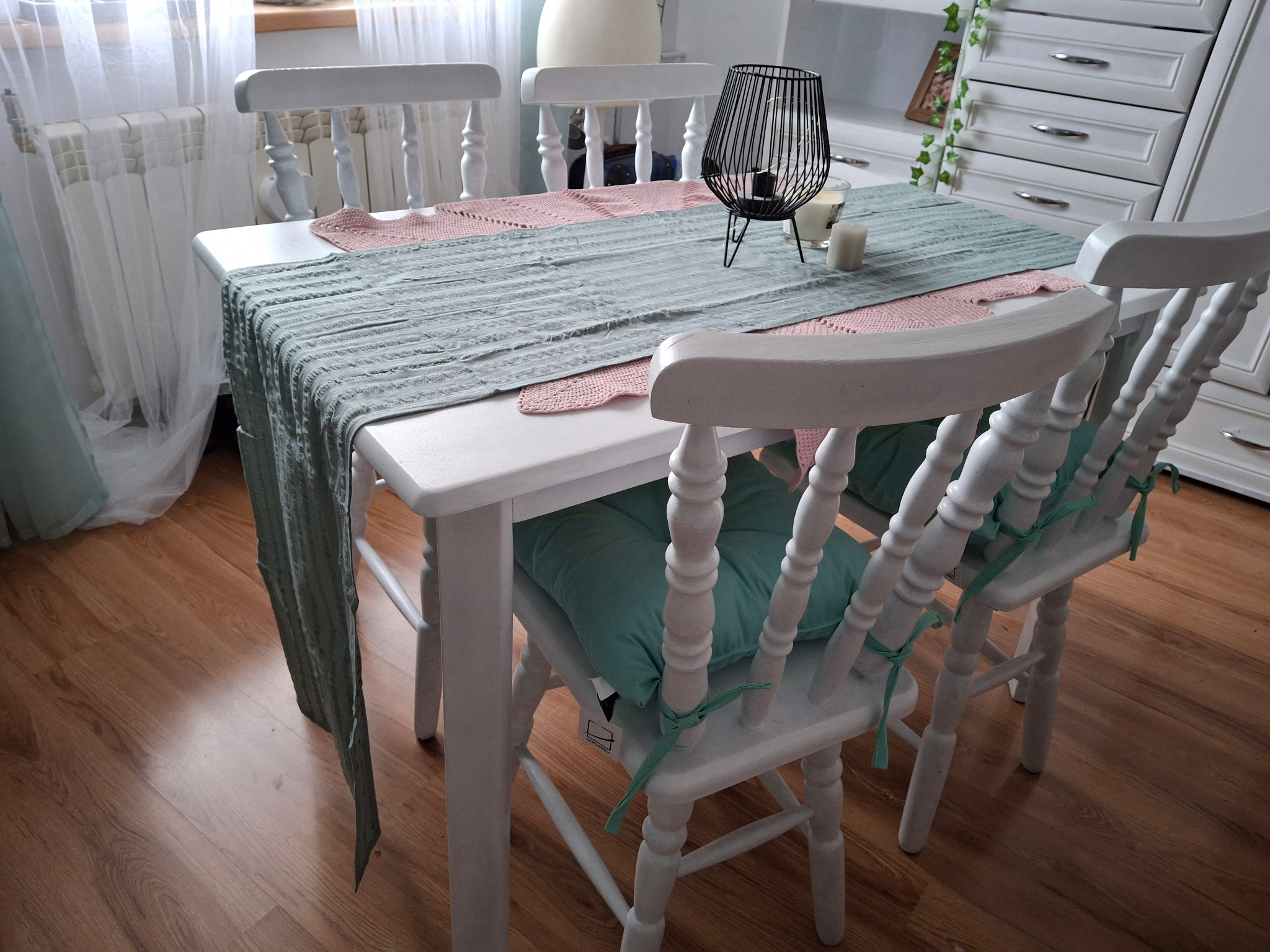 Biały, drewniany stół z trzema krzesłami w stylu rustykalnym, nakryty szarym bieżnikiem i różowymi serwetkami, dekorowany czarnym lampionem i świecami, na tle białej komody i drewnianej podłogi.