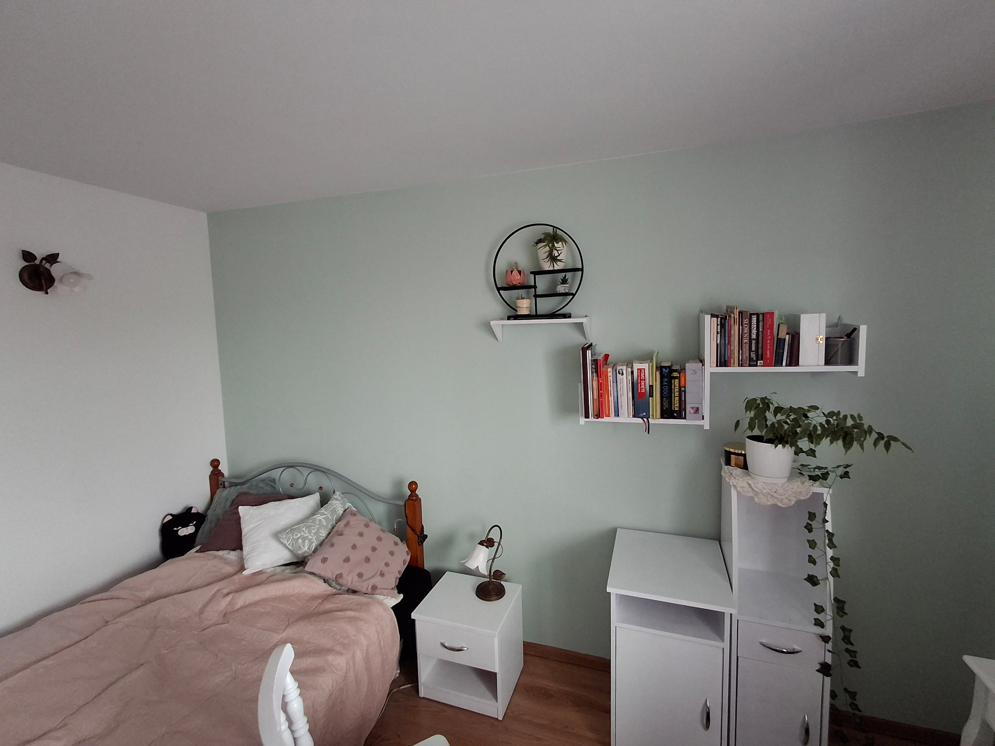 Pokój w pastelowych kolorach z łóżkiem z metalową ramą, białymi szafkami nocnymi i półkami na ścianie, dekorowany roślinami doniczkowymi i lampką nocną w stylu retro.