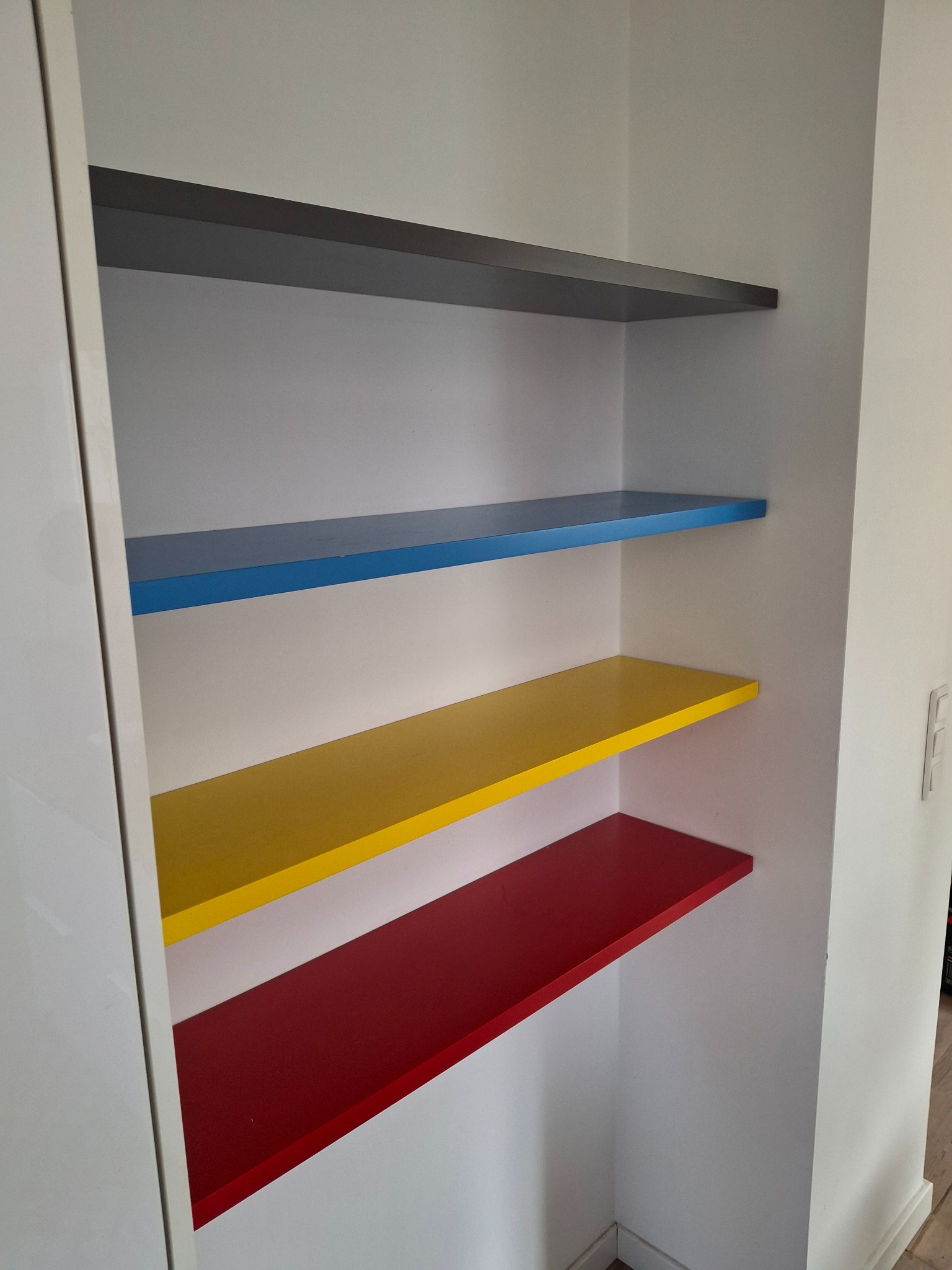 Wnęka z zamontowanymi półkami w kolorach: szarym, niebieskim, żółtym i czerwonym, wbudowana w białą szafę.
