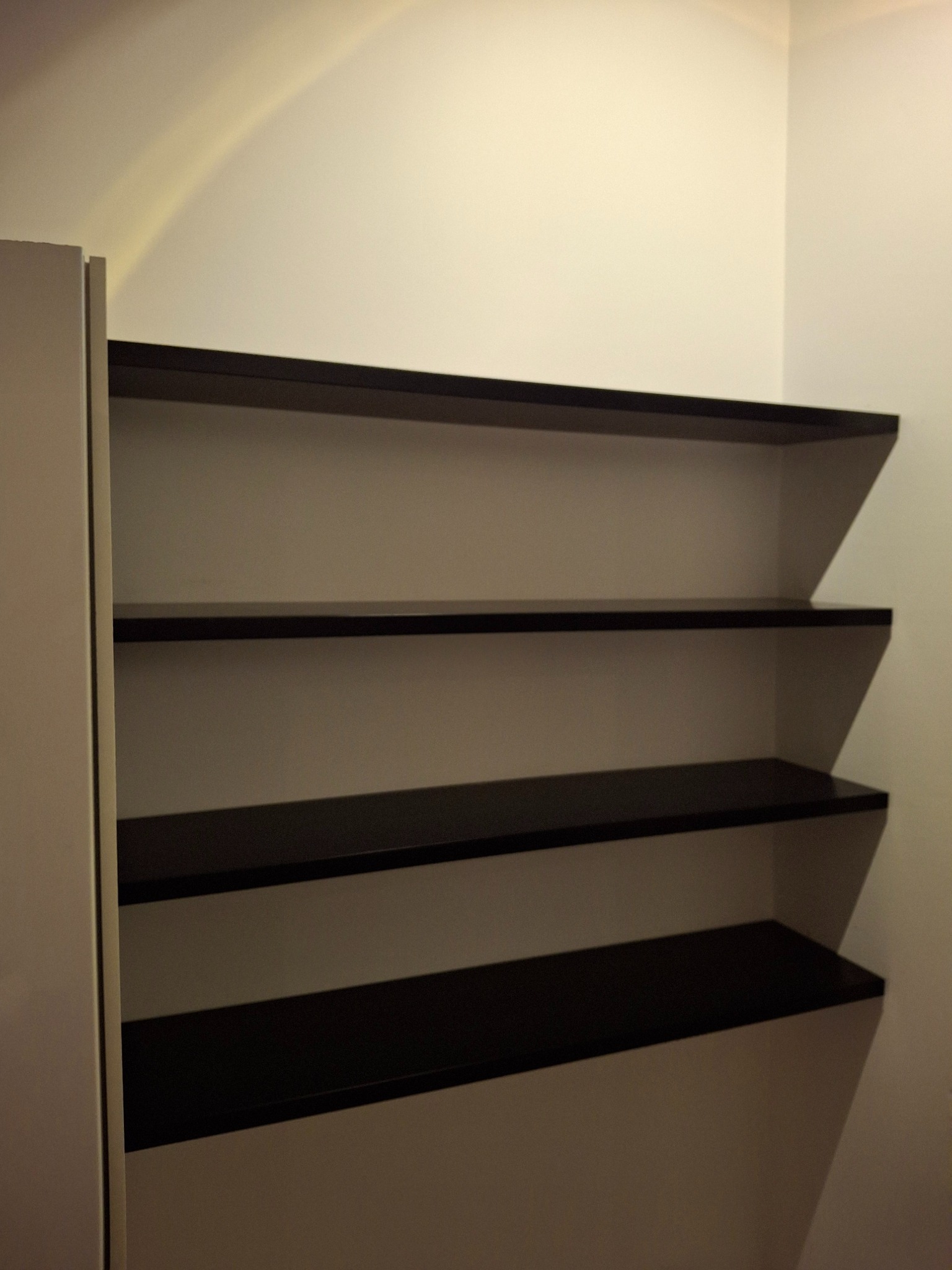 Cztery puste, ciemne półki zamontowane na białej ścianie w rogu pokoju, widok z boku szafy.