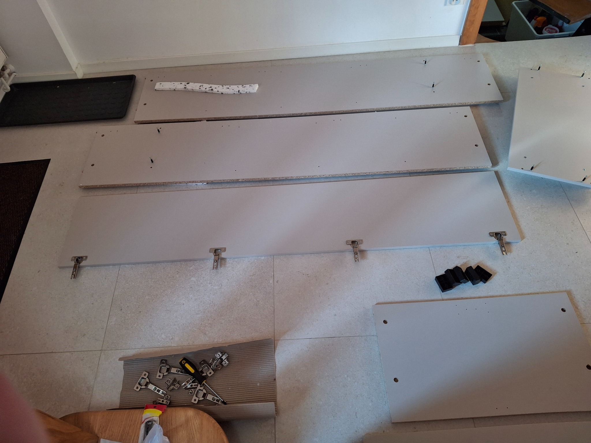 Rozłożone elementy mebli z zawiasami i narzędziami na podłodze z płytek, przygotowane do montażu.