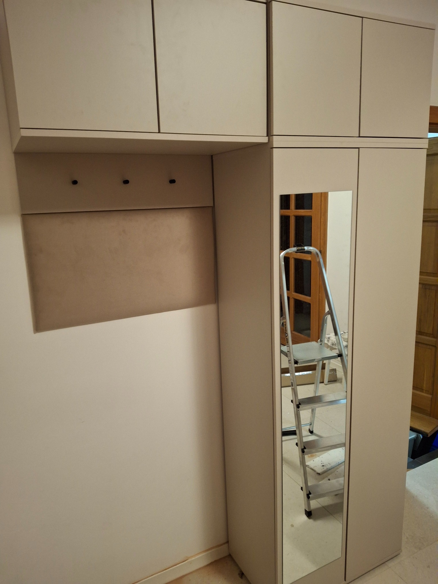 Montaż szafy z lustrem i górnymi szafkami, widoczna drabina w odbiciu lustra i panel z trzema wieszakami.