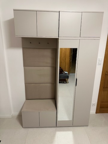 Beżowy zestaw mebli do przedpokoju z szafkami górnymi, lustrem, wieszakami i siedziskiem z tapicerowanym panelem na ścianie.