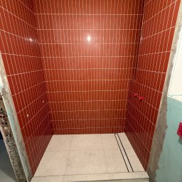 Roman Piddubnyi - Wnętrze kabiny prysznicowej w trakcie wykańczania, z pionowo ułożonymi, błyszczącymi, ceglastoczerwonymi płytkami na ścianach i jasnobeżową, lastrykową posadzką z odpływem liniowym. Widoczne...