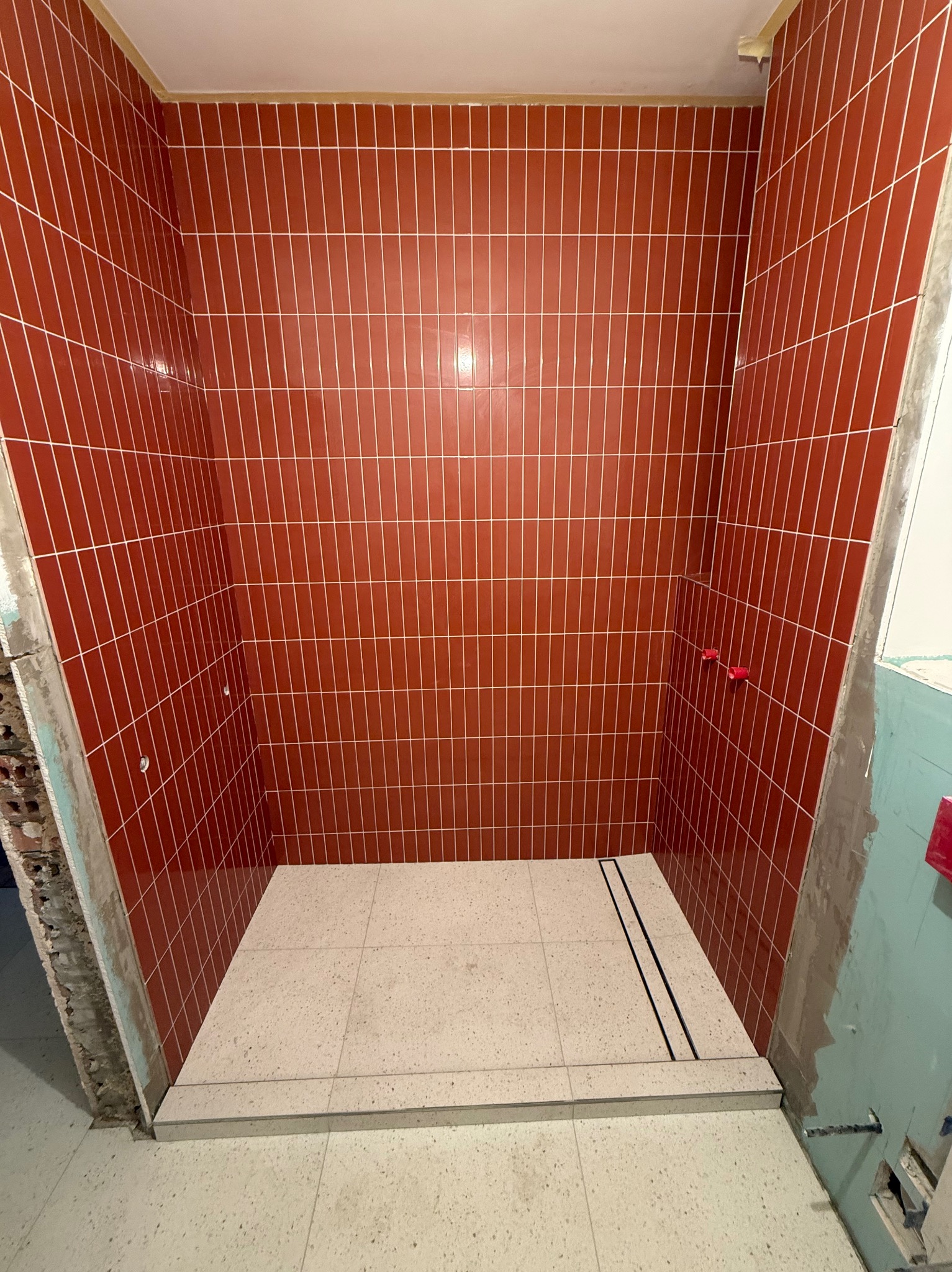 Wnętrze kabiny prysznicowej w trakcie wykańczania, z pionowo ułożonymi, błyszczącymi, ceglastoczerwonymi płytkami na ścianach i jasnobeżową, lastrykową posadzką z odpływem liniowym. Widoczne...