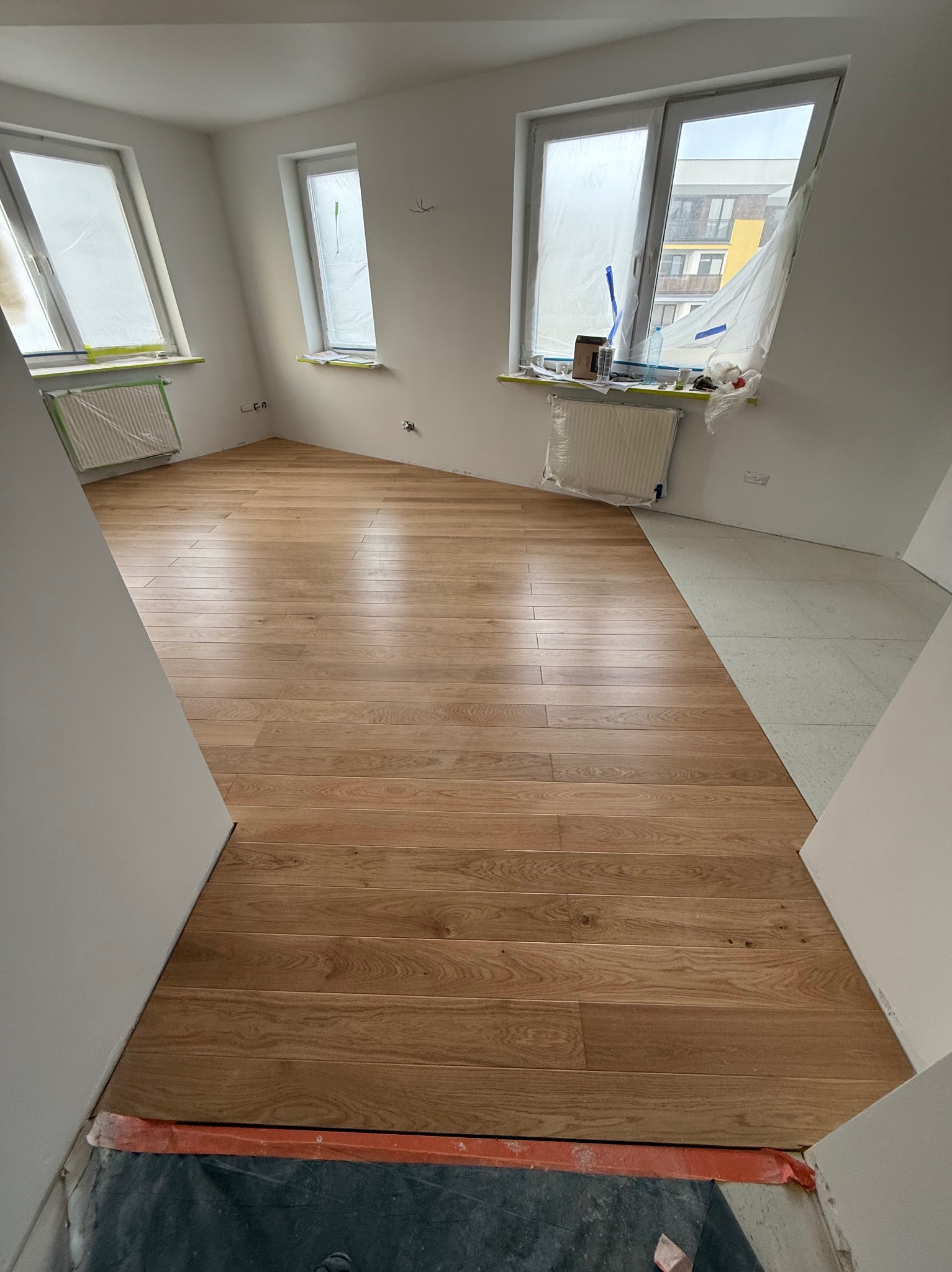 Ukończone układanie jasnych paneli podłogowych w pokoju z trzema oknami zabezpieczonymi folią malarską, widoczne niedociągnięcia przy ścianach i fragment niezakończonej podłogi.