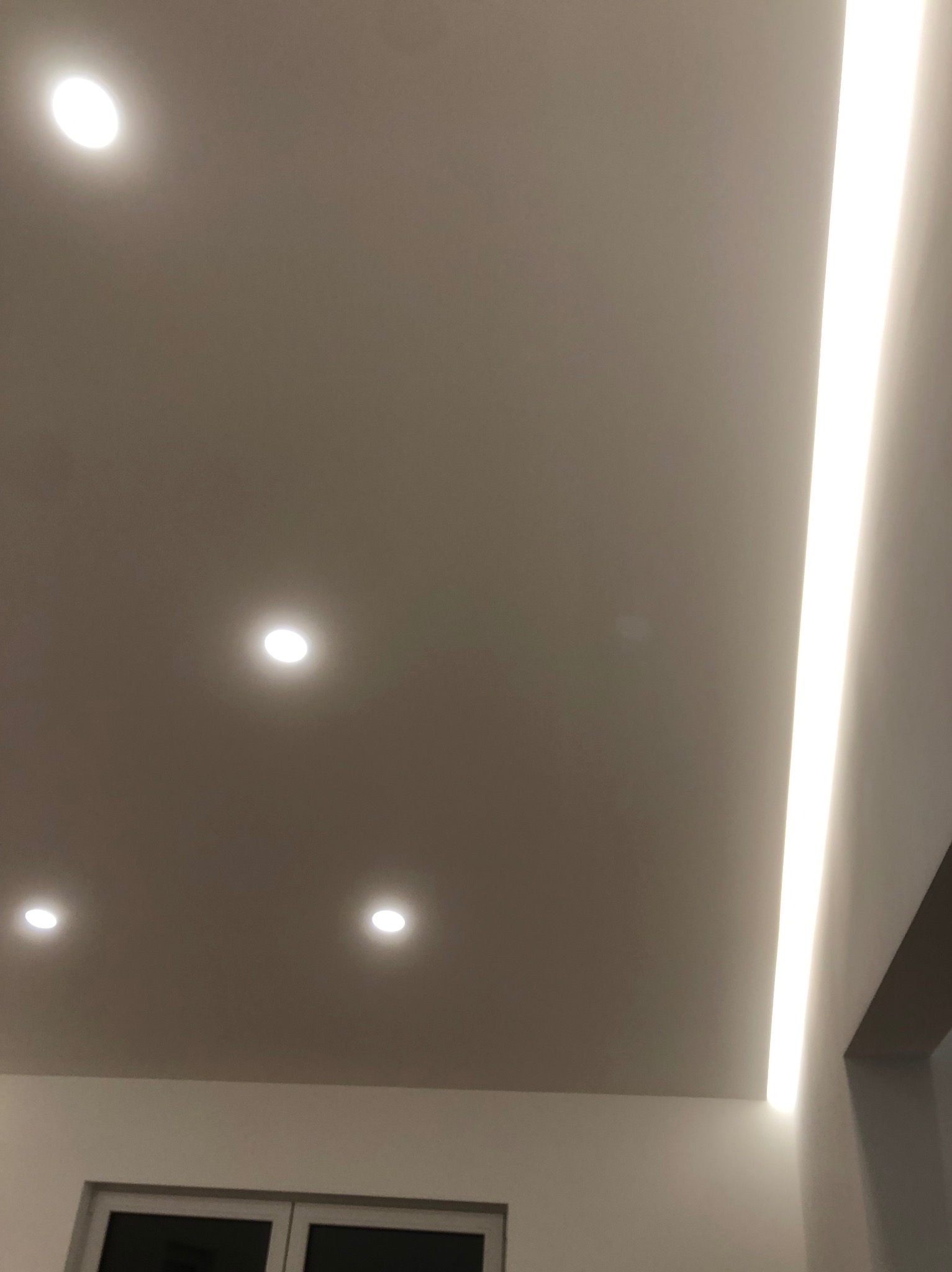 Nowoczesny sufit podwieszany z liniowym oświetleniem LED i punktowymi lampami halogenowymi, widoczne okno w dolnej części kadru.