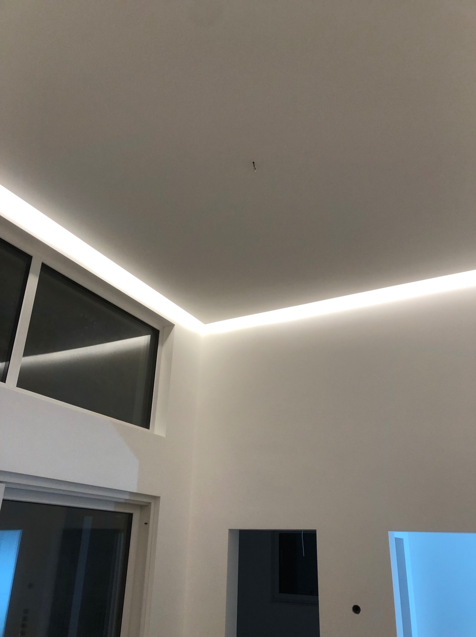 Nowoczesne oświetlenie LED w białym wnętrzu, listwy zamontowane pod sufitem i nad oknem, widoczne okno oraz otwór drzwiowy w ścianie, surowy stan wykończenia.