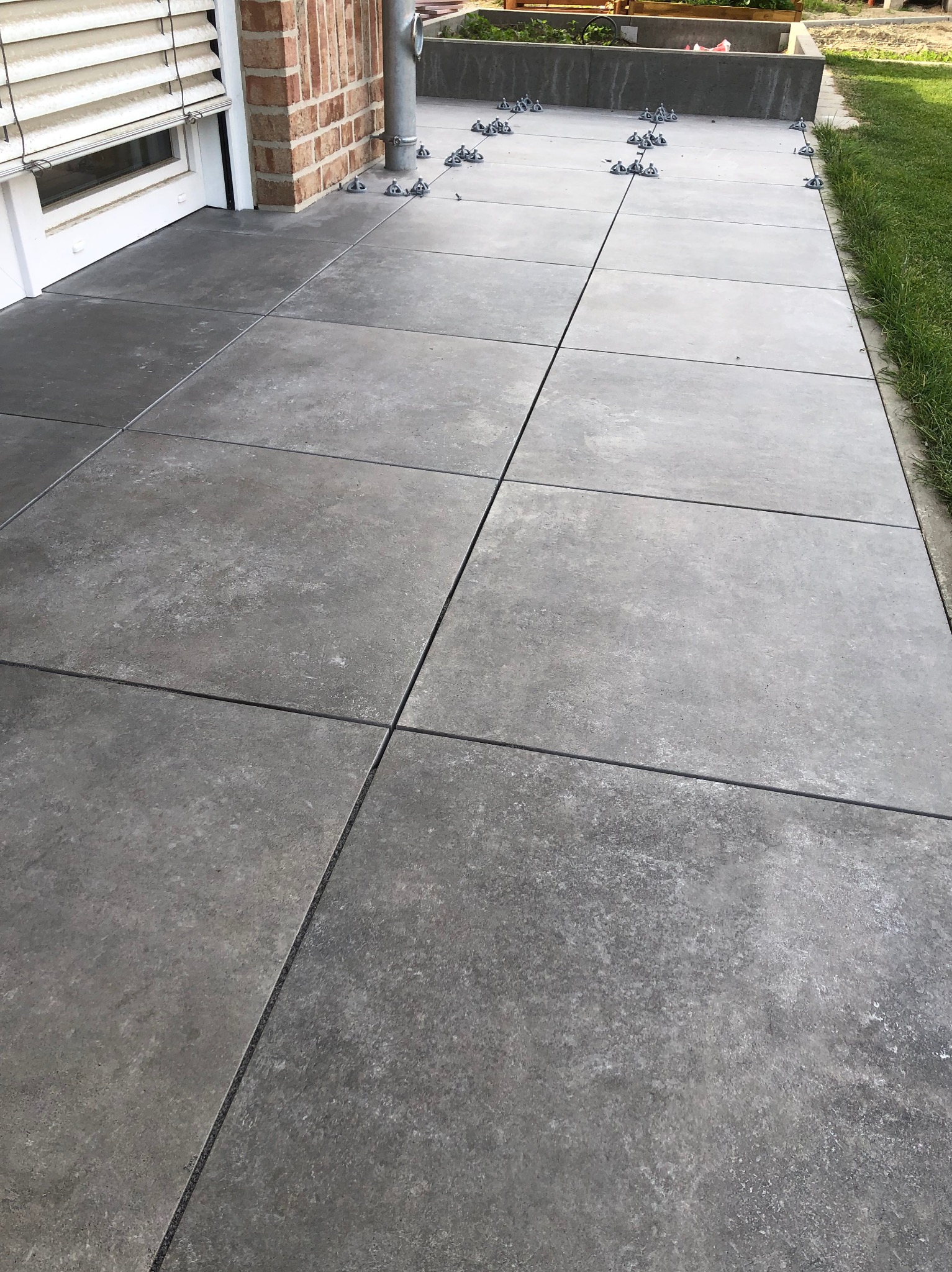 Szare płytki tarasowe ułożone na zewnątrz budynku, z widocznymi klipsami poziomującymi w trakcie montażu, ceglana ściana budynku z rynną w tle.