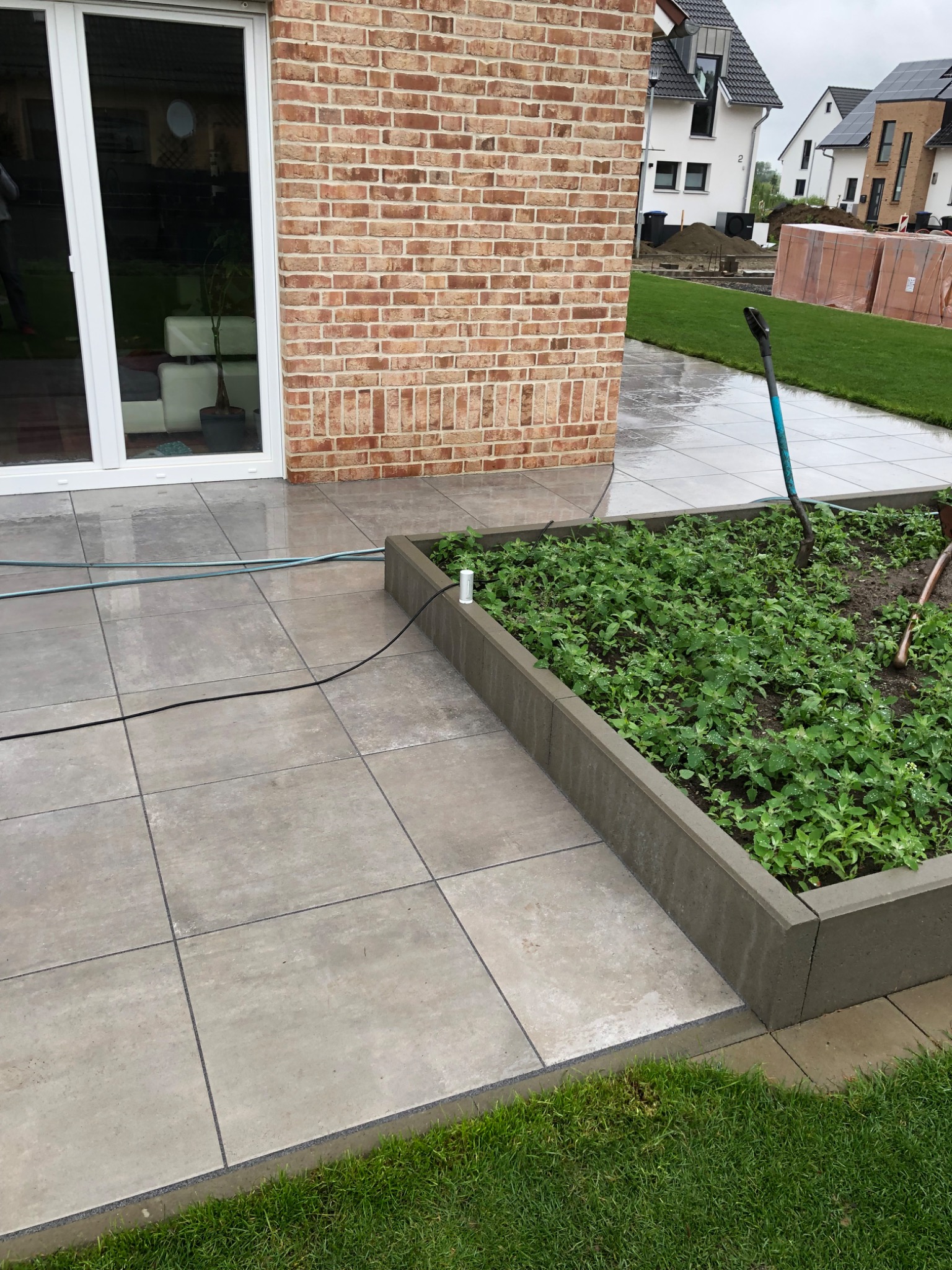Wyłożony szarymi płytami taras z ceglaną ścianą domu i podwyższoną grządką z zielenią, widoczne narzędzia ogrodnicze i trawnik w tle.