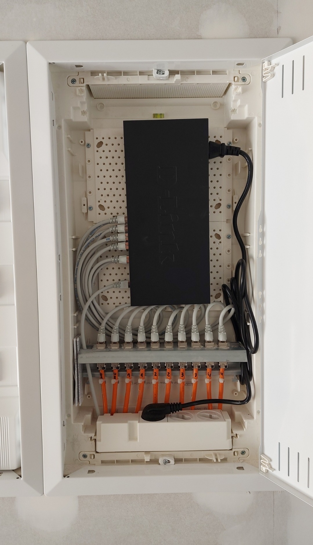 Rozdzielnica elektryczna w obudowie z widocznymi przewodami ethernetowymi, listwą zaciskową z ponumerowanymi złączami i routerem D-Link z podłączonym przewodem zasilającym.