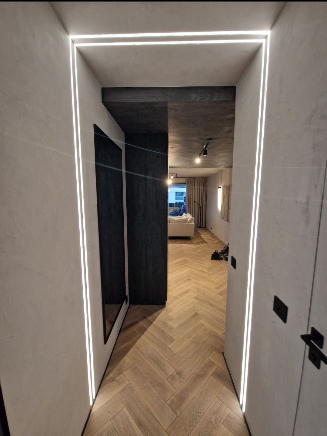 Nowoczesny korytarz z oświetleniem LED wzdłuż ścian, parkiet ułożony w jodełkę, widok na salon z jasną sofą.