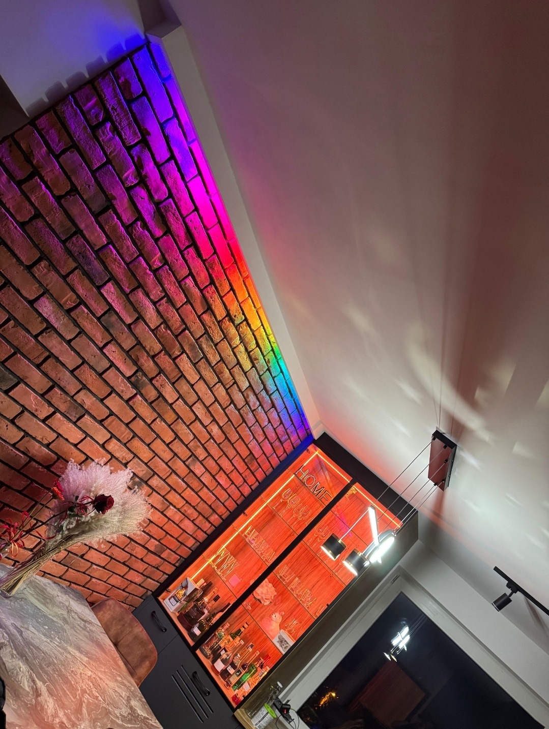 Ceglana ściana z podświetleniem LED w kolorach tęczy, oświetlona witryna z alkoholem i szklankami, nowoczesne oświetlenie wiszące.