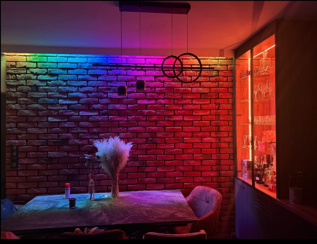 Jadalnia z ceglaną ścianą podświetloną wielokolorowym światłem LED, nowoczesna lampa wisząca z okrągłymi kloszami, przeszklona witryna z oświetleniem i zastawą.