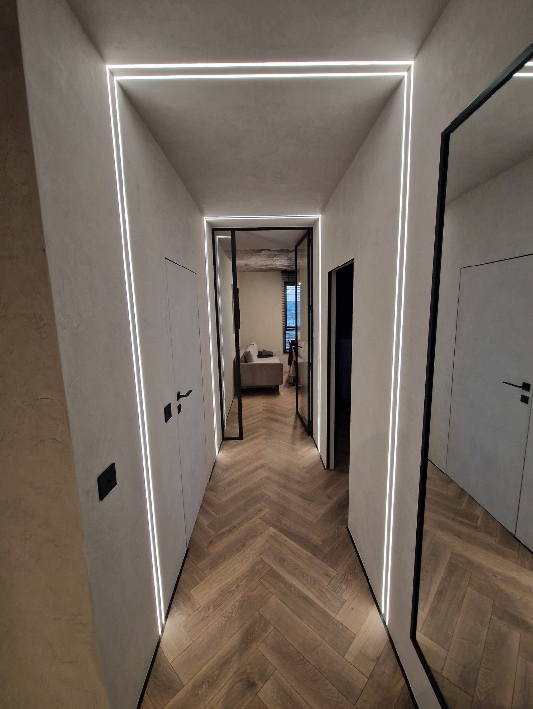 Nowoczesny korytarz z oświetleniem liniowym LED wzdłuż ścian i sufitu, parkiet ułożony w jodełkę, czarne detale drzwi i ramy lustra.