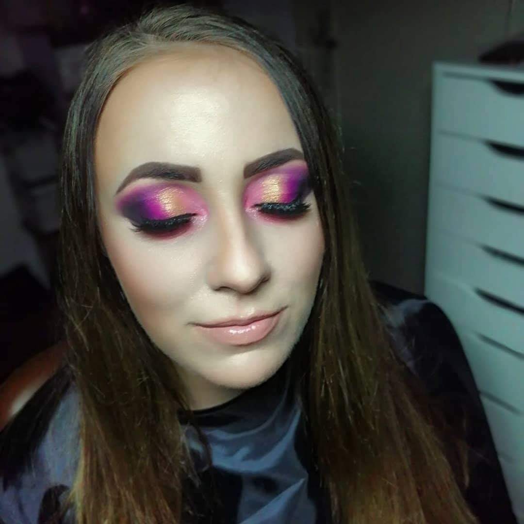 Portret kobiety z intensywnym makijażem oczu w odcieniach fioletu, różu i złota, z długimi sztucznymi rzęsami, delikatnym błyszczykiem na ustach i ciemnym peleryną fryzjerską.