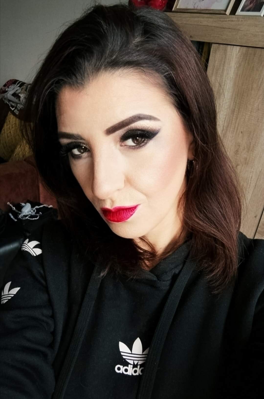 Portret kobiety z intensywnym makijażem oczu typu smoky eye i jaskrawoczerwoną szminką, ubrana w czarną bluzę z kapturem marki Adidas.